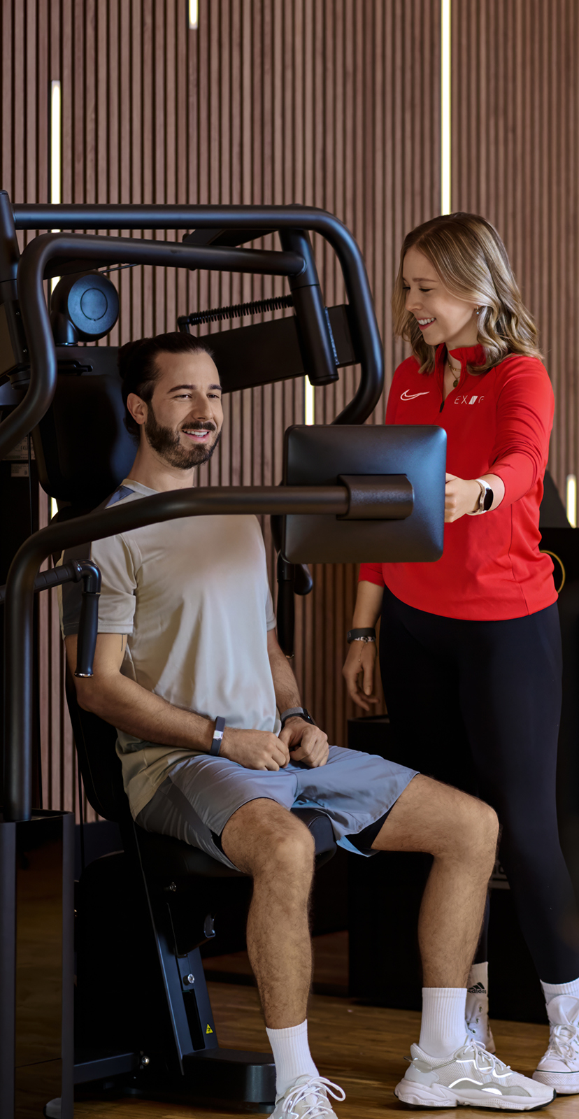 Ein Mann sitzt auf einem Kraftgerät, während eine Trainerin im roten Exice-Fitness-Shirt ihn im modernen Fitnessstudio anleitet.